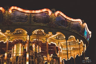 A brightly lit carousel with ornate decorations and glowing lights against a dark background. Several people, including children, can be seen enjoying the ride. The carousel features detailed paintings and gold accents, creating a whimsical and nostalgic atmosphere.