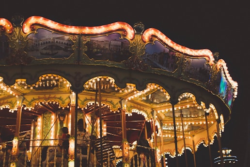 A brightly lit carousel with ornate decorations and glowing lights against a dark background. Several people, including children, can be seen enjoying the ride. The carousel features detailed paintings and gold accents, creating a whimsical and nostalgic atmosphere.