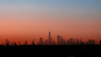 A city skyline silhouetted against a vibrant sunset with shades of orange and blue blending seamlessly across the sky. The tallest building is prominently outlined, with other structures gradually descending in height toward the edges of the image. The contrast between the dark foreground and the colorful sky creates a striking visual effect.