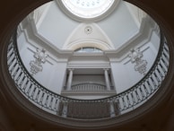 An elegant interior view of a building featuring a circular balcony structure with ornate railings. The ceiling is a domed design with a central circular skylight, allowing natural light to illuminate the space. The walls and architectural details are in a monochromatic color scheme, adding a classical and sophisticated atmosphere.