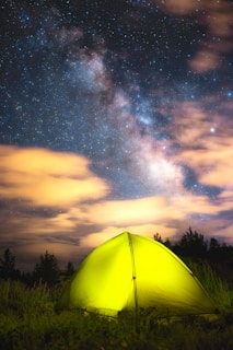 A vibrant yellow tent set in a grassy field under a stunning night sky filled with stars and the Milky Way. Wispy orange clouds are illuminated against the deep blue and black backdrop of the sky. The surrounding area features silhouetted trees, creating a serene and isolated camping scene.