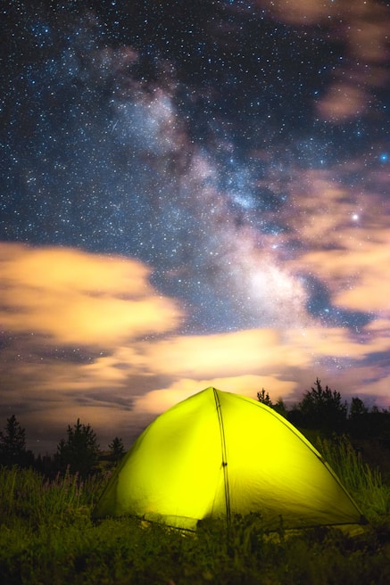 A vibrant yellow tent set in a grassy field under a stunning night sky filled with stars and the Milky Way. Wispy orange clouds are illuminated against the deep blue and black backdrop of the sky. The surrounding area features silhouetted trees, creating a serene and isolated camping scene.