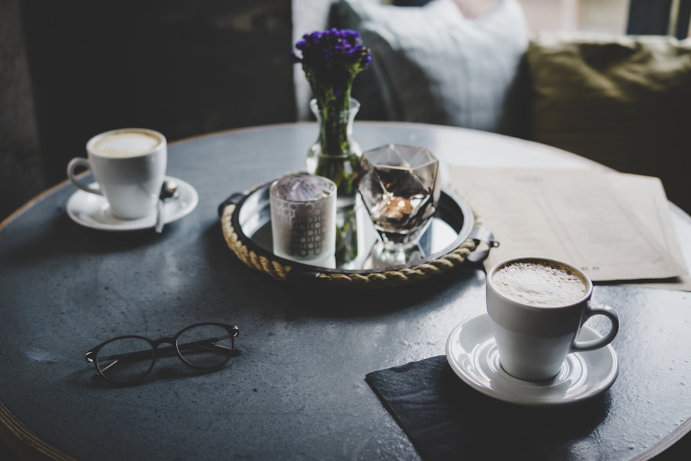 round tray with flower vase between two white ceramic teacups above table