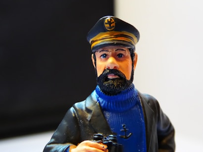 A figurine of a bearded man wearing a blue turtleneck sweater and a black captain's hat with yellow accents. The figure holds a pair of binoculars in one hand and has an anchor emblem on the sweater.
