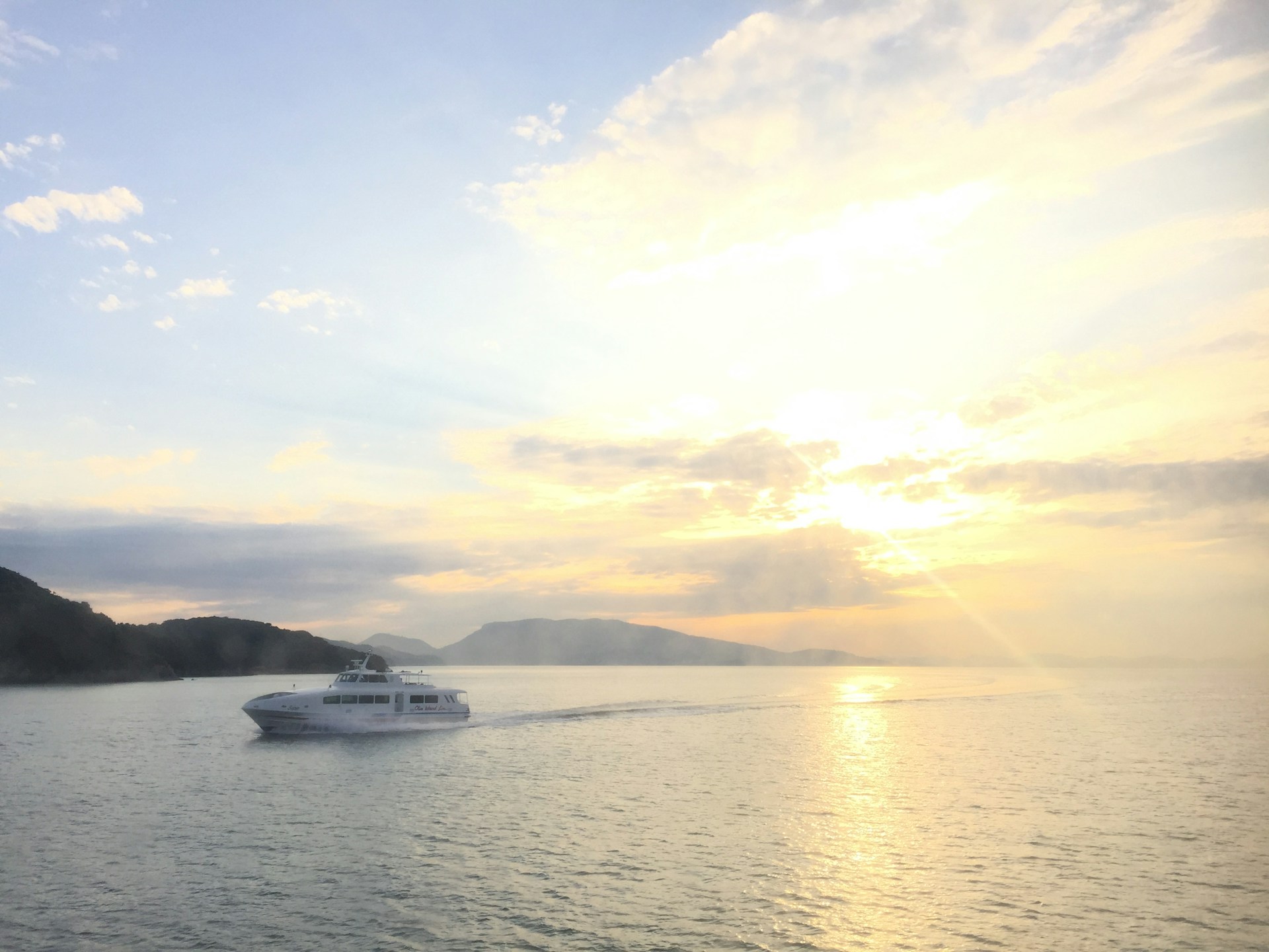 A large white yacht is gliding smoothly on a calm sea, with the sun setting or rising in the background. The sky is filled with soft clouds, and the light from the sun creates a warm, reflective glow on the water. Distant landmasses are visible on the horizon, adding depth to the scenic view.