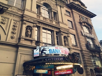 An ornate, historic building featuring detailed architectural elements such as columns and sculptures is shown. The facade includes large windows and decorative stonework. Below the structure, a bright marquee sign advertises a comedy play titled 'The Comedy About A Bank Robbery' at the Criterion Theatre, with vibrant colors and lights.