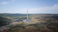 A large industrial complex with a tall smokestack is situated in a sprawling, hilly landscape. There are various buildings and infrastructure surrounding the main facility. The landscape is mostly green with some brown patches, indicating a mixture of vegetation and barren land. In the foreground, there are small clusters of buildings or houses.