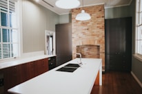 A modern kitchen with a large white island featuring an integrated sink and chrome faucet. The background showcases an exposed brick wall with an unused fireplace, flanked by dark cabinetry. The room has wooden floors and light-colored walls with large windows allowing natural light in.
