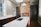A modern kitchen with a large white island featuring an integrated sink and chrome faucet. The background showcases an exposed brick wall with an unused fireplace, flanked by dark cabinetry. The room has wooden floors and light-colored walls with large windows allowing natural light in.