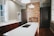 A modern kitchen with a large white island featuring an integrated sink and chrome faucet. The background showcases an exposed brick wall with an unused fireplace, flanked by dark cabinetry. The room has wooden floors and light-colored walls with large windows allowing natural light in.