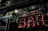 A vintage-style illuminated sign spelling out 'BAR' with light bulbs encased in metallic letters. The setting includes industrial elements like metal beams and spotlight fixtures. Reflections and glass panels are visible, suggesting an urban or cityscape environment.
