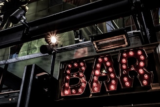 A vintage-style illuminated sign spelling out 'BAR' with light bulbs encased in metallic letters. The setting includes industrial elements like metal beams and spotlight fixtures. Reflections and glass panels are visible, suggesting an urban or cityscape environment.
