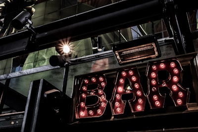 A vintage-style illuminated sign spelling out 'BAR' with light bulbs encased in metallic letters. The setting includes industrial elements like metal beams and spotlight fixtures. Reflections and glass panels are visible, suggesting an urban or cityscape environment.