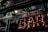 A vintage-style illuminated sign spelling out 'BAR' with light bulbs encased in metallic letters. The setting includes industrial elements like metal beams and spotlight fixtures. Reflections and glass panels are visible, suggesting an urban or cityscape environment.