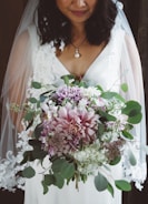A bride in a white dress holding a lush bouquet featuring an array of flowers including pink and purple blossoms, surrounded by rich green leaves. The dress has lace details and the bride wears a delicate pearl necklace. Her dark hair gently falls on her shoulders, partially covered by a sheer veil.