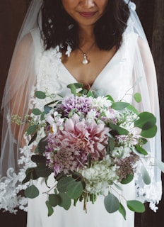 A bride in a white dress holding a lush bouquet featuring an array of flowers including pink and purple blossoms, surrounded by rich green leaves. The dress has lace details and the bride wears a delicate pearl necklace. Her dark hair gently falls on her shoulders, partially covered by a sheer veil.