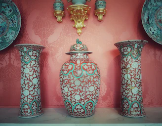 A symmetrically arranged set of decorative vases with intricate floral designs is displayed against an ornate red background. At the center is a large lidded vase, flanked by two tall vases. All vases have a red and blue pattern with white flowers. Surrounding the central vases are wall-mounted decorative plates and small ornate jars, adding to the elegance of the display.