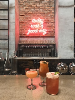 Three colorful cocktails with garnishes are placed on a bar counter with a brick wall in the background. A neon sign on the wall reads 'today was a good day'. Glass bottles and draft beer taps are also visible.