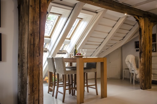A cozy attic space with slanted ceilings and exposed wooden beams. The room features a wooden high table with two upholstered chairs. On the table, there is a bottle of wine, two wine glasses, and a small bowl of fruit. Natural light streams in through large skylights, illuminating the interior. A fluffy white chair is placed near a corner, adding to the warm and inviting atmosphere.