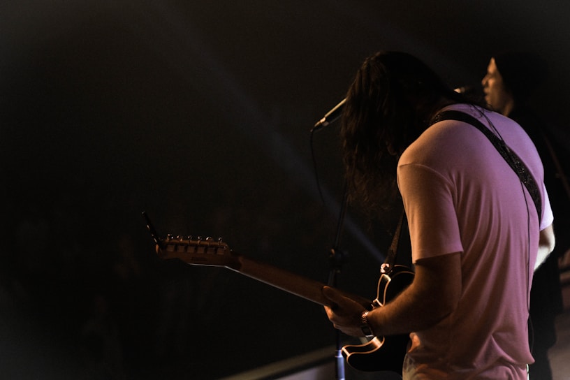A musician with long hair wearing a casual t-shirt is playing an electric guitar on stage. The lighting is dim, with spotlight accents, creating a moody atmosphere. Another person in a hoodie stands in the background near a microphone.