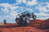 A rugged off-road vehicle navigating over red rocky terrain under a bright blue sky with scattered clouds. A person wearing a green cap is taking photos of the scene, capturing the adventure.