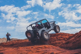 A rugged off-road vehicle navigating over red rocky terrain under a bright blue sky with scattered clouds. A person wearing a green cap is taking photos of the scene, capturing the adventure.