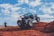 A rugged off-road vehicle navigating over red rocky terrain under a bright blue sky with scattered clouds. A person wearing a green cap is taking photos of the scene, capturing the adventure.