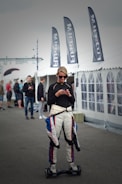 A person wearing a racing suit is standing on a hoverboard while using a smartphone. Several black flags with white lettering are visible behind, as well as a large white tent. Other people are in the background, suggesting a busy outdoor setting.