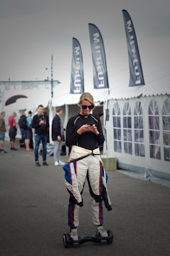 A person wearing a racing suit is standing on a hoverboard while using a smartphone. Several black flags with white lettering are visible behind, as well as a large white tent. Other people are in the background, suggesting a busy outdoor setting.