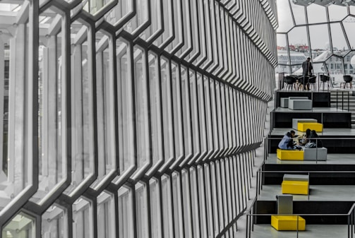 A modern architectural interior with a large, geometric glass wall casting soft light across a series of tiered seating areas. People are seated at different levels, engaged in conversation or using electronic devices. The seating is modular, with a mix of gray and bright yellow cushioned seating providing a pop of color against the predominantly gray and glass environment.