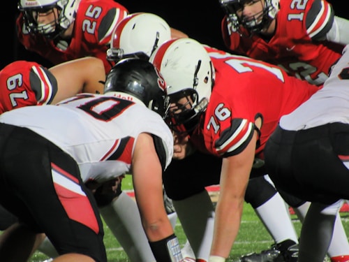 Players lined up for a football game, with two teams facing each other at the line of scrimmage. Players are wearing protective gear including helmets and jerseys with numbers, indicating an intense moment in the game under bright lights.