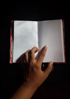 A hand is flipping through an open notebook with blank pages. The notebook has a colorful cover and is placed against a dark background.