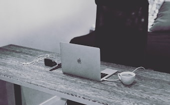 A monochrome setting with a wooden desk holding a laptop, a pair of earphones, a small camera, and a cup of coffee. The background features a dimly lit sofa with a decorative pillow.