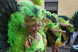 Close-up of a smiling woman wearing a bright feathered headdress and glittery makeup at a street carnival.