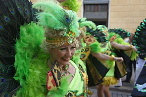 Close-up of a smiling woman wearing a bright feathered headdress and glittery makeup at a street carnival.