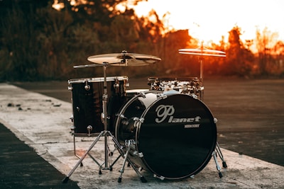 A drum kit is set up on a deserted road with a vibrant sunset in the background. The sky is painted with warm orange hues, contrasting with the dark silhouette of trees. The drum set includes a bass drum, two cymbals, and a tom-tom, all featuring a glossy finish.