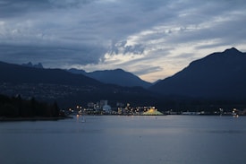 A tranquil waterfront scene at dusk featuring a cityscape with illuminated buildings against a backdrop of mountains. The sky is overcast with layered clouds adding depth to the evening atmosphere. Lights reflect gently on the water, enhancing the calm ambiance.