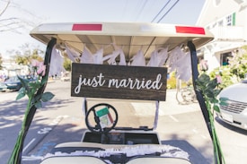 A golf cart is decorated with white streamers and pink flowers. A wooden sign with the words 'just married' is displayed on the back. The scene is outdoors on a sunny street with houses and cars in the background.