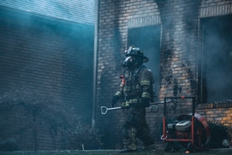 A firefighter in full gear, including a helmet and breathing apparatus, walks in front of a smoke-damaged brick building. The atmosphere is filled with haze and there's a portable ventilation fan on the ground next to the firefighter.