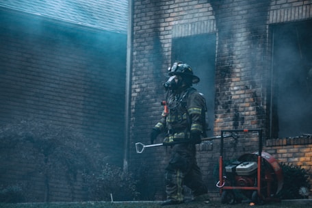A firefighter in full gear, including a helmet and breathing apparatus, walks in front of a smoke-damaged brick building. The atmosphere is filled with haze and there's a portable ventilation fan on the ground next to the firefighter.