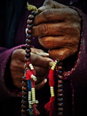 Hands gripping a string of prayer beads adorned with colorful tassels and metallic accents. The beads are held tightly, showing the details of the aged, textured hands and the vibrant threads.
