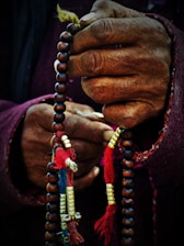 Hands gripping a string of prayer beads adorned with colorful tassels and metallic accents. The beads are held tightly, showing the details of the aged, textured hands and the vibrant threads.