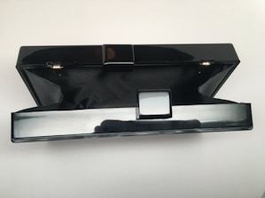A black, rectangular hard-shell clutch purse with a polished finish is open, revealing a black fabric interior. The exterior has a glossy surface with a simple clasp mechanism in the center. The background is plain white, accentuating the sleek design of the clutch.