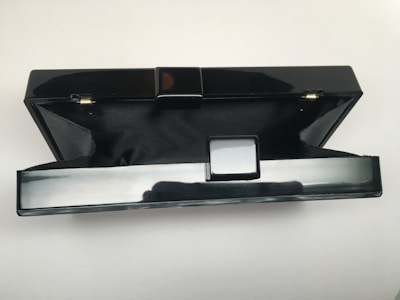 A black, rectangular hard-shell clutch purse with a polished finish is open, revealing a black fabric interior. The exterior has a glossy surface with a simple clasp mechanism in the center. The background is plain white, accentuating the sleek design of the clutch.
