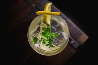 A top-down view of a refreshing drink in a glass with ice cubes, garnished with lemon slices and a sprig of parsley, set against a dark background.