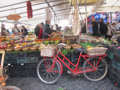 A bustling market scene with vibrant fruit and vegetable displays. Stalls are filled with various produce in baskets, including bananas, peppers, and pomegranates. A red bicycle with a wicker basket is parked in the foreground. Shoppers and vendors interact under a large canopy.