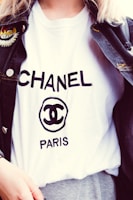 A person is wearing a white T-shirt with the word CHANEL and the logo prominently displayed in black text. A dark denim jacket with an embroidered patch on the shoulder is layered over the T-shirt.