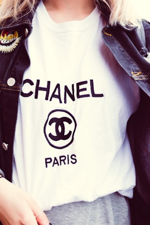 A person is wearing a white T-shirt with the word CHANEL and the logo prominently displayed in black text. A dark denim jacket with an embroidered patch on the shoulder is layered over the T-shirt.