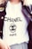 A person is wearing a white T-shirt with the word CHANEL and the logo prominently displayed in black text. A dark denim jacket with an embroidered patch on the shoulder is layered over the T-shirt.