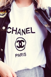 A person is wearing a white T-shirt with the word CHANEL and the logo prominently displayed in black text. A dark denim jacket with an embroidered patch on the shoulder is layered over the T-shirt.